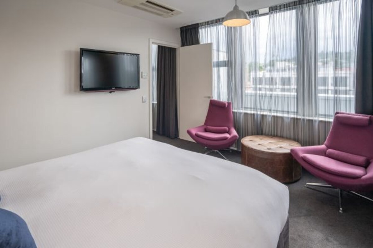Una habitación de hotel con una cama grande, dos sillones rosados, un reposapiés redondo, un televisor de pantalla plana montado en la pared y un gran ventanal con cortinas traslúcidas.