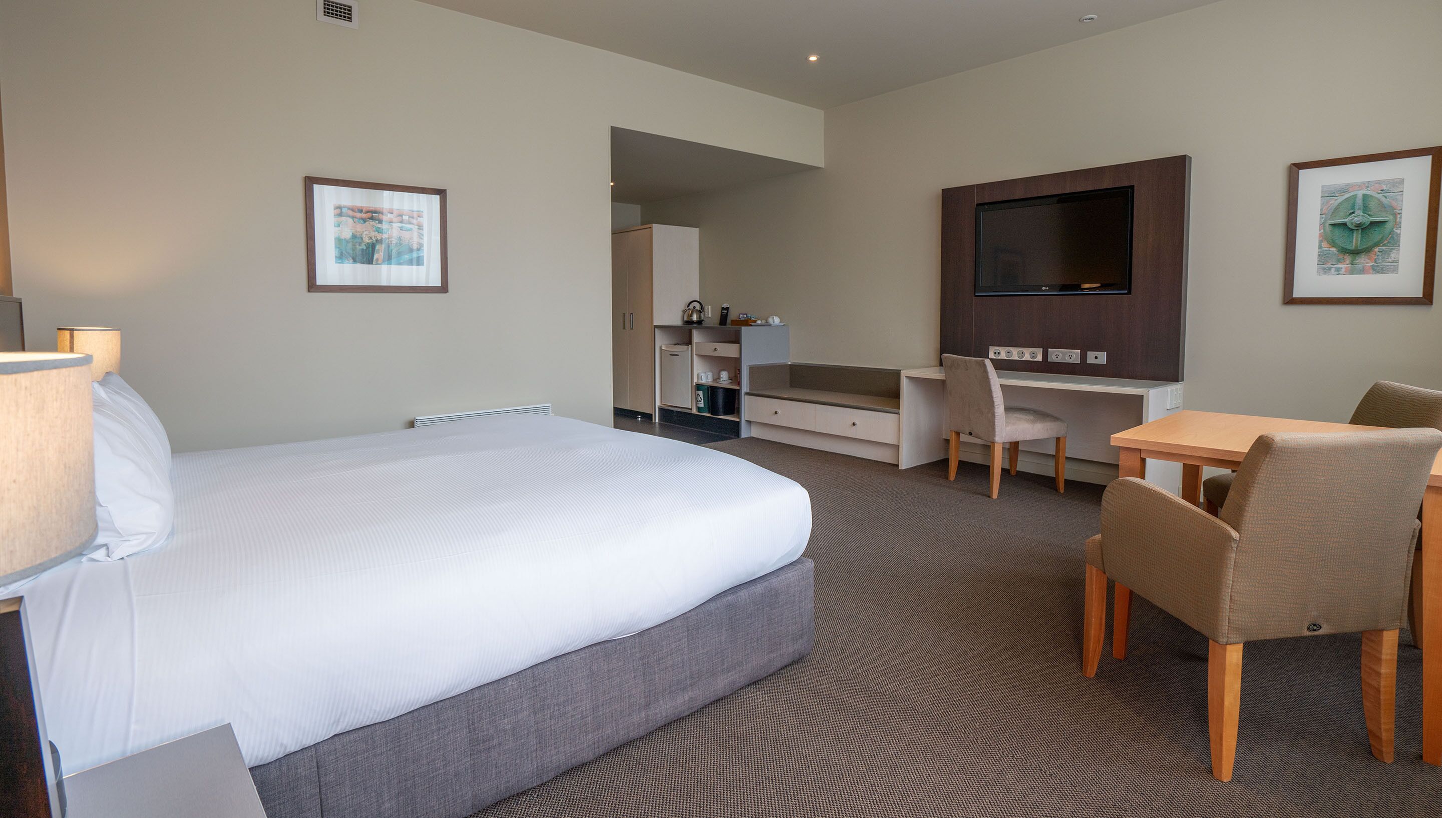 A hotel room with a large bed, a desk, a chair, a TV, and a small kitchenette.