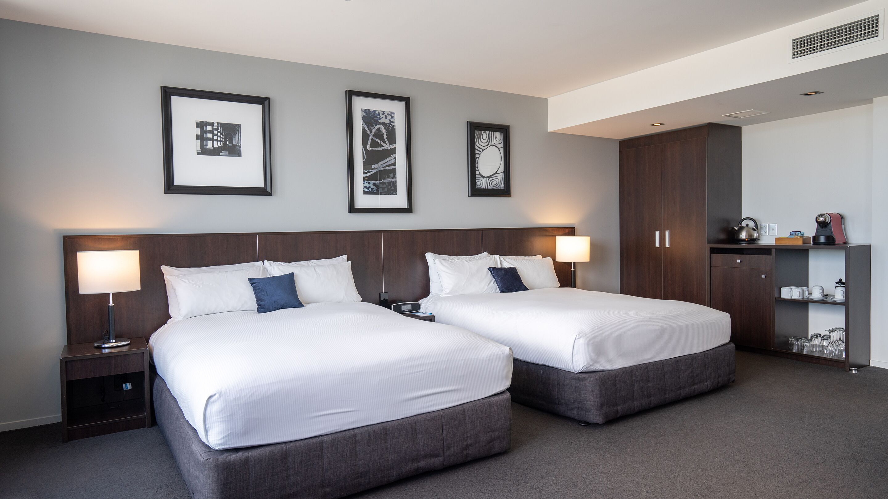 A hotel room with two beds, a wardrobe, a shelf with glasses, and framed pictures on the wall.