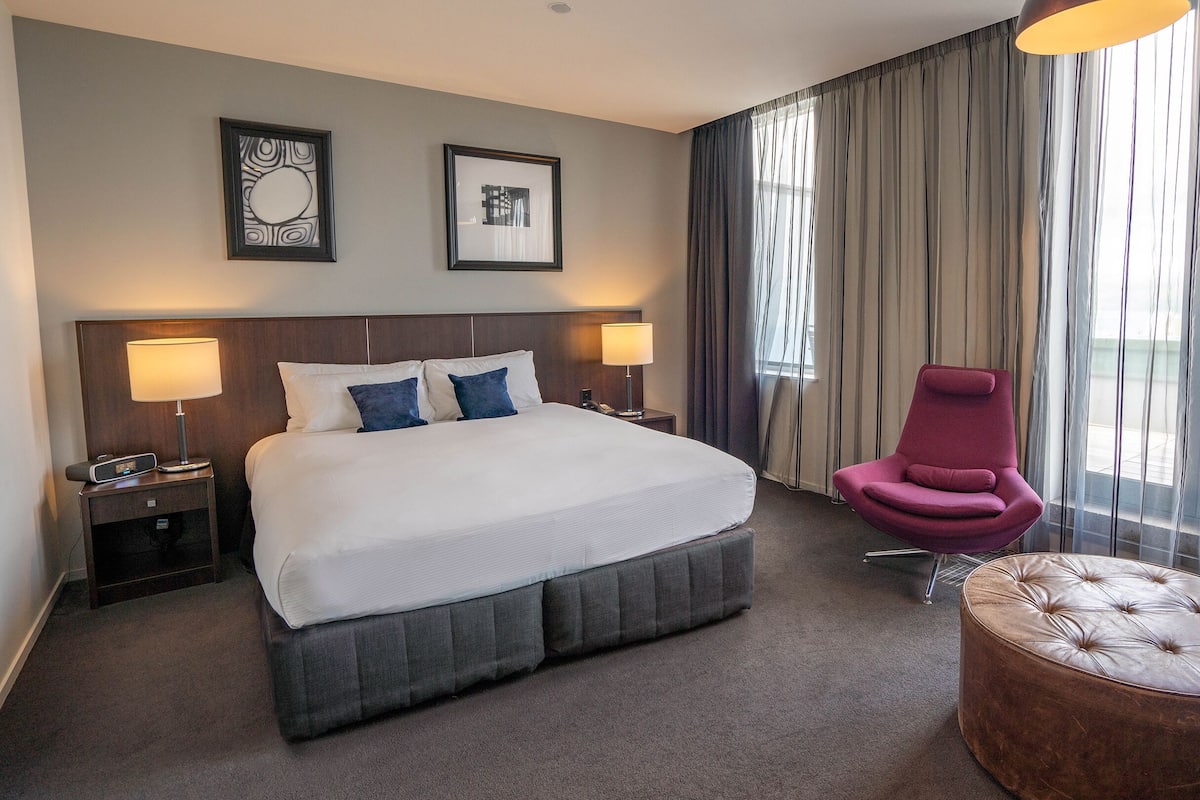 A hotel room with a large bed, two bedside lamps, a nightstand, a red armchair, a round ottoman, and a window with curtains.
