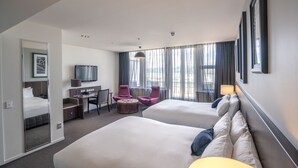 Habitación de hotel con dos camas, un escritorio, una silla, un televisor y un ventanal grande con cortinas.