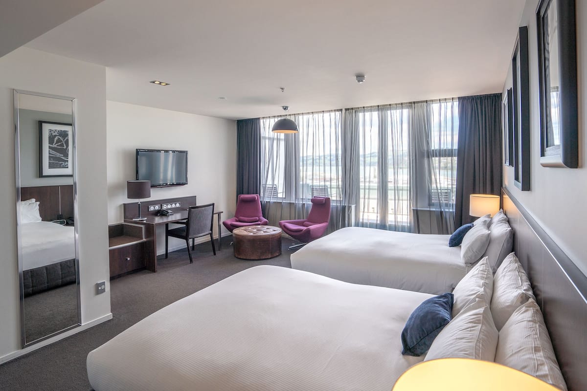A hotel room with two beds, a desk, a chair, a TV, and a large window with curtains.
