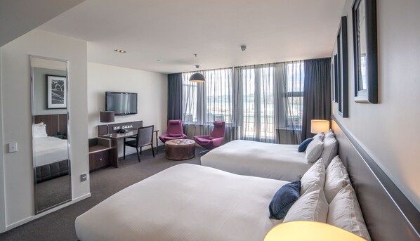 A hotel room with two beds, a desk, a chair, a TV, and a large window with curtains.