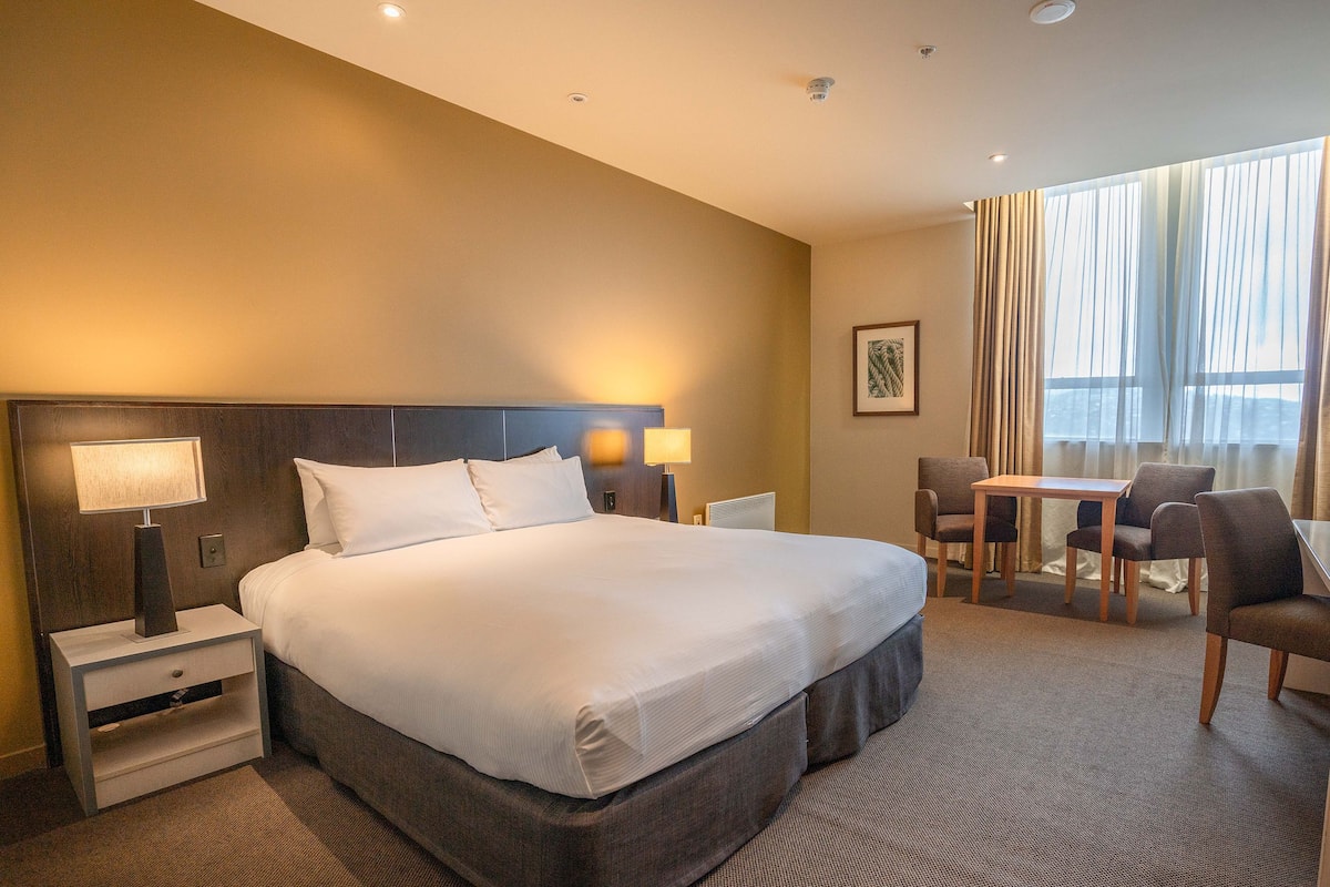 A hotel room with a large bed, bedside lamps, a desk with two chairs, and a window with curtains.