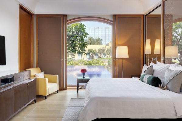 A modern hotel room with a large bed, a seating area with a sofa, a flat-screen TV, and a view of a pool and greenery outside.