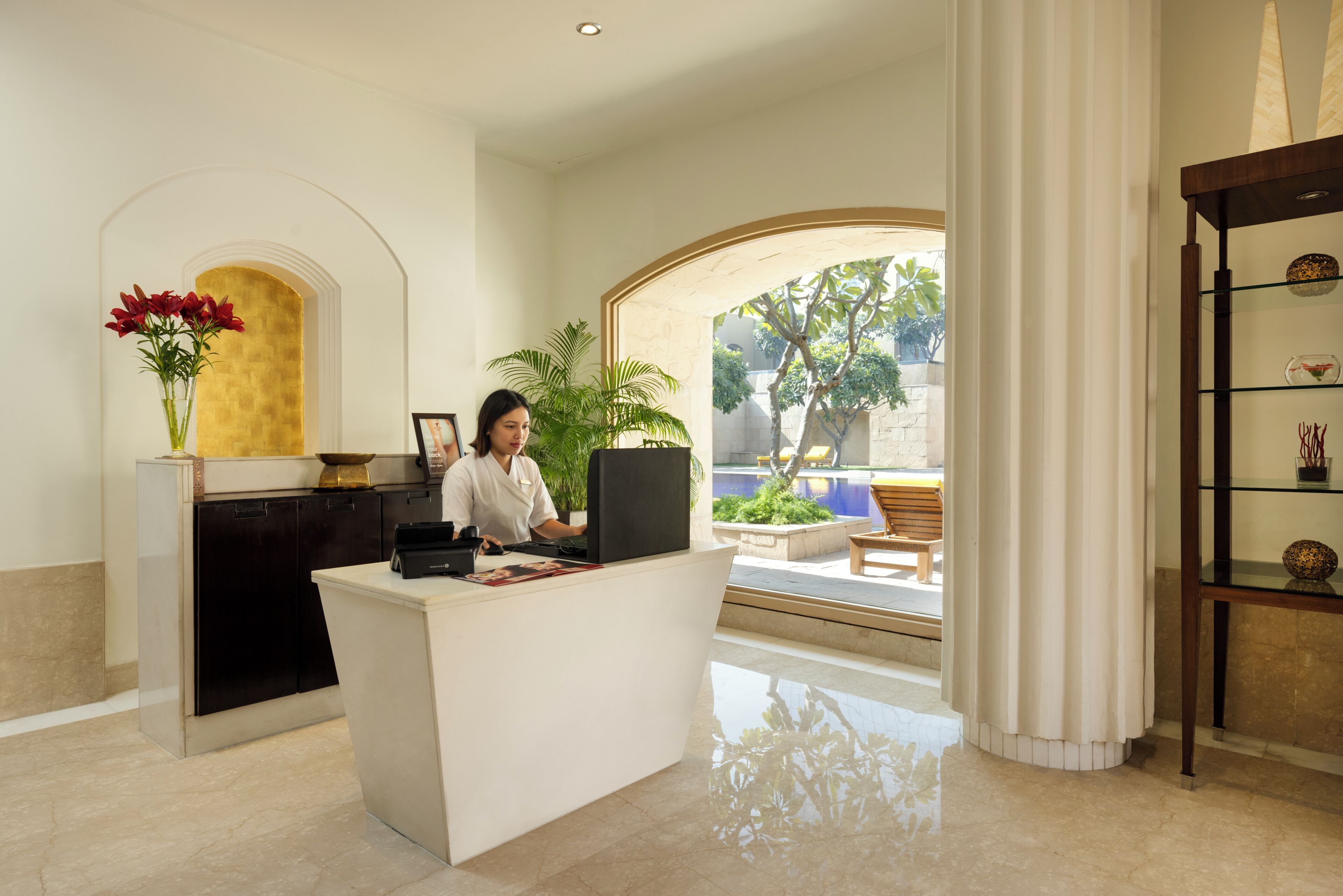 A reception desk with a computer, a vase with red flowers, and a view of an outdoor seating area.