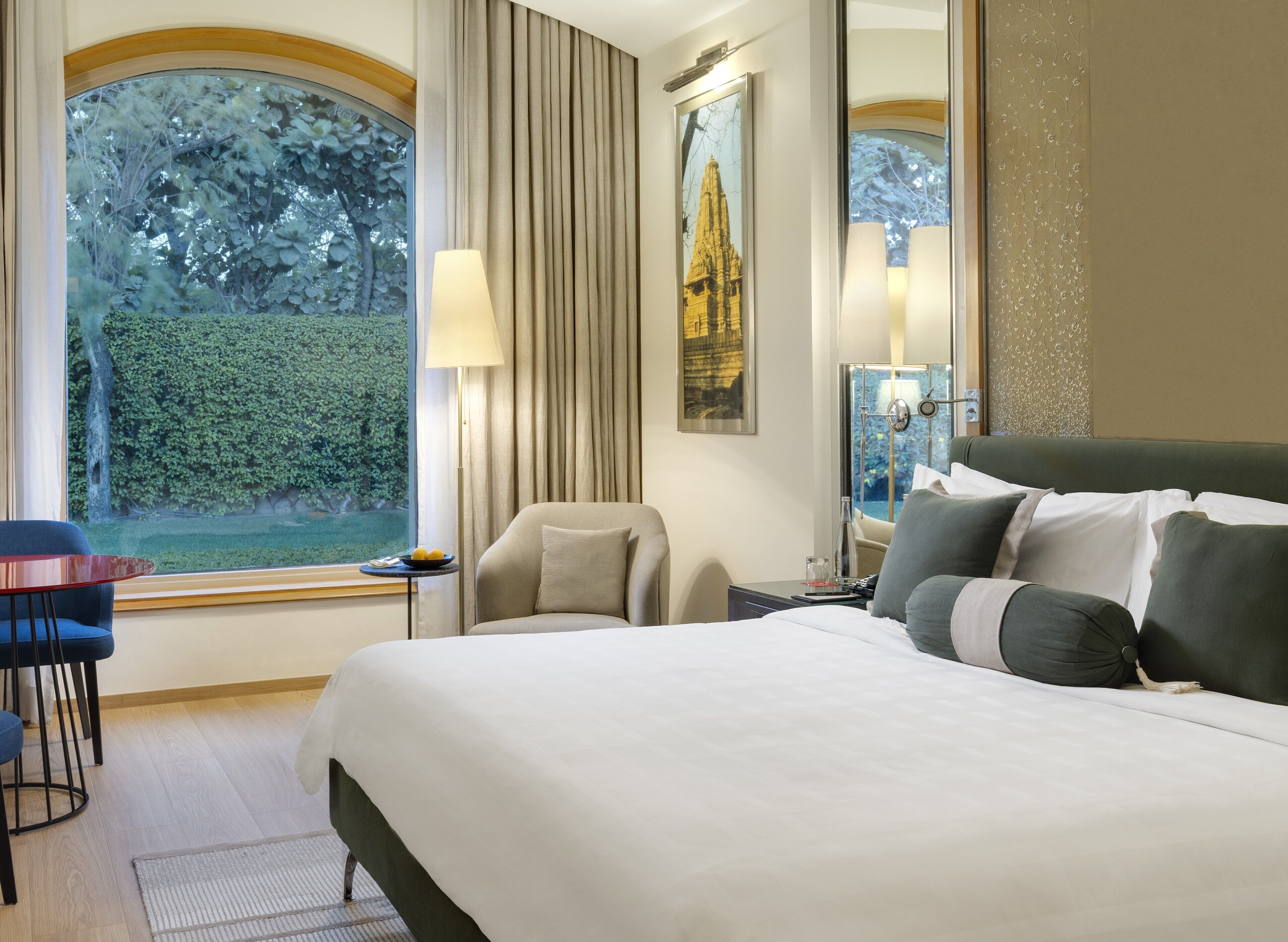 A hotel room with a large bed, a chair, a small table with a lamp, and a view of a garden through a large window.