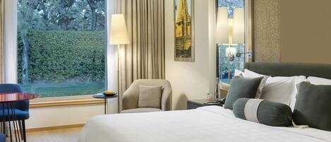 A hotel room with a large bed, a chair, a small table with a lamp, and a view of a garden through a large window.