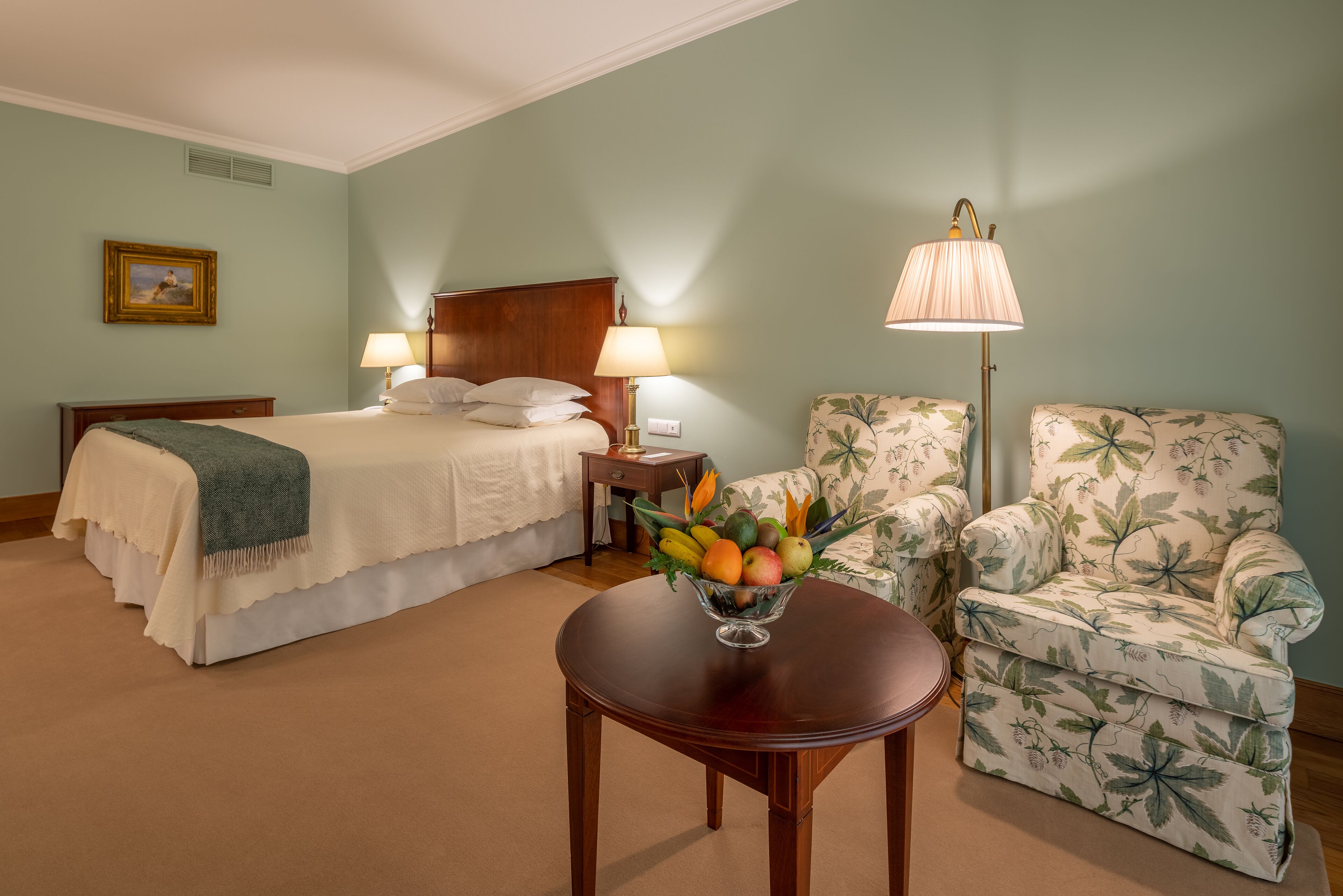 A hotel room with a bed, two bedside lamps, a floral armchair, a round wooden table, and a fruit bowl.