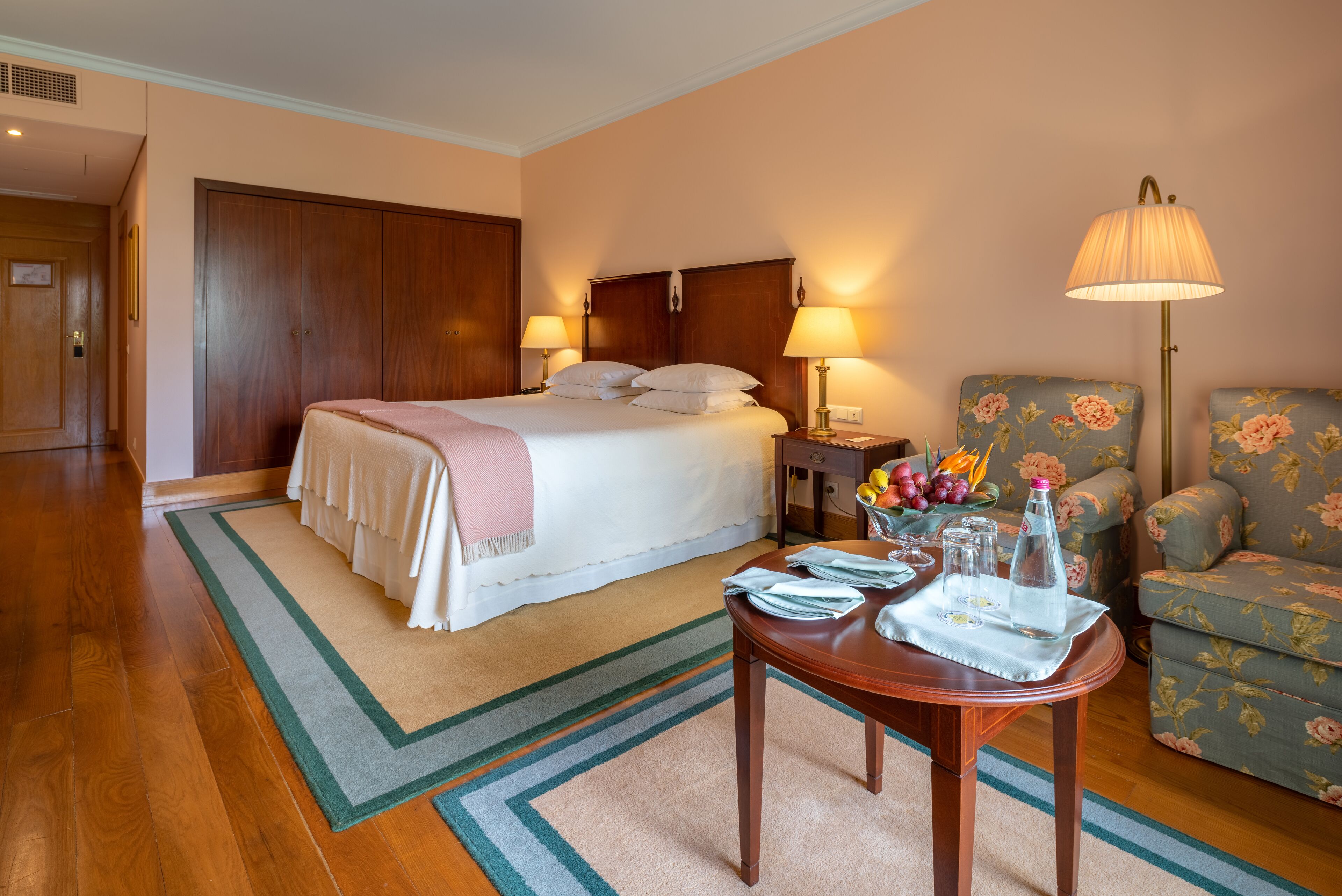 Habitación de hotel con cama, lámparas de noche, una mesa con botella de agua, vasos y una bandeja, además de un sillón con estampado floral.