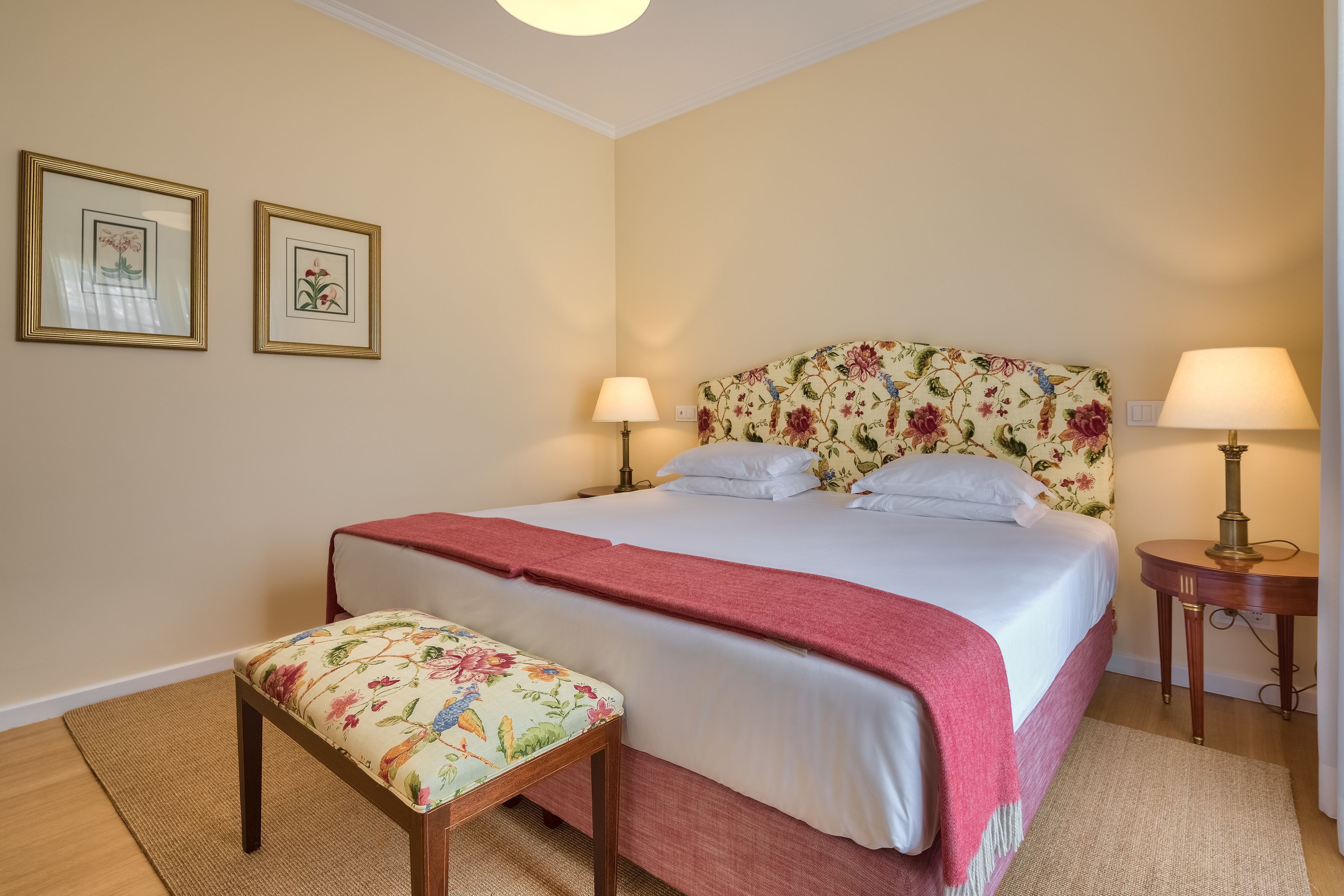 A neatly made bed with a floral headboard, two bedside lamps, and a bench with a floral cushion.