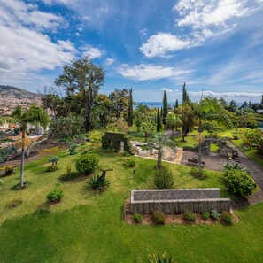 Un exuberante jardín con una estructura de piedra, diversas especies de árboles y una vista de un horizonte urbano lejano.