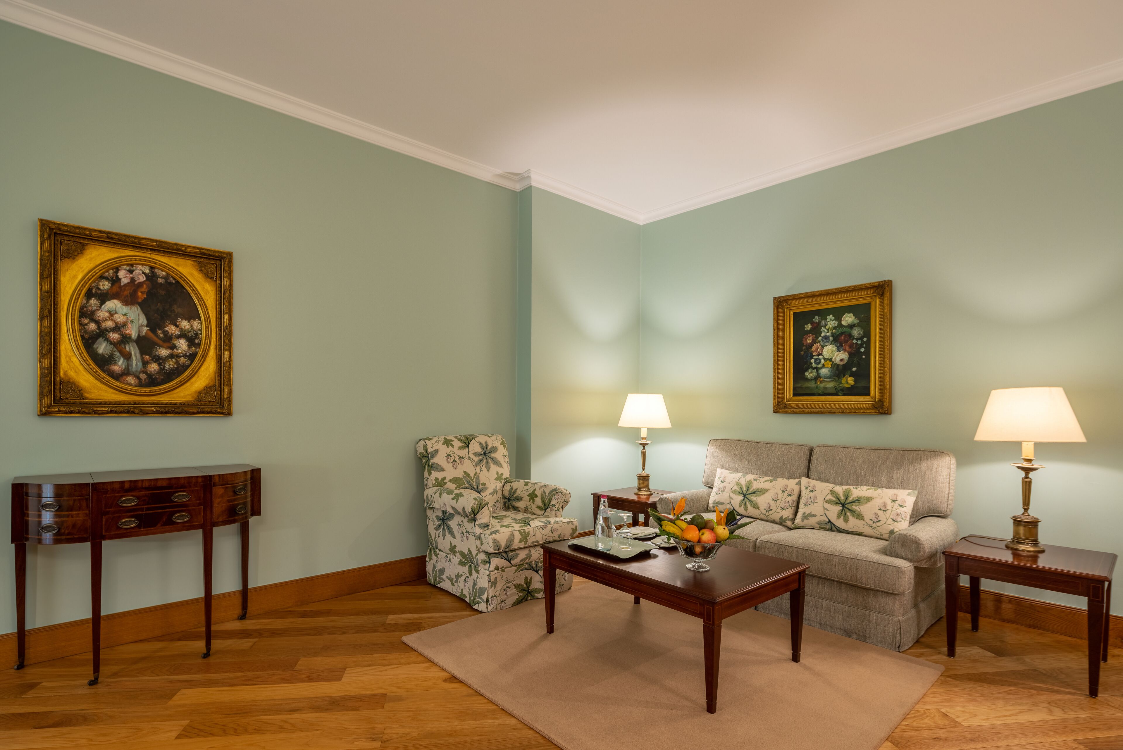 A living room with a sofa, armchairs, a coffee table, and two lamps. There are two paintings on the walls.