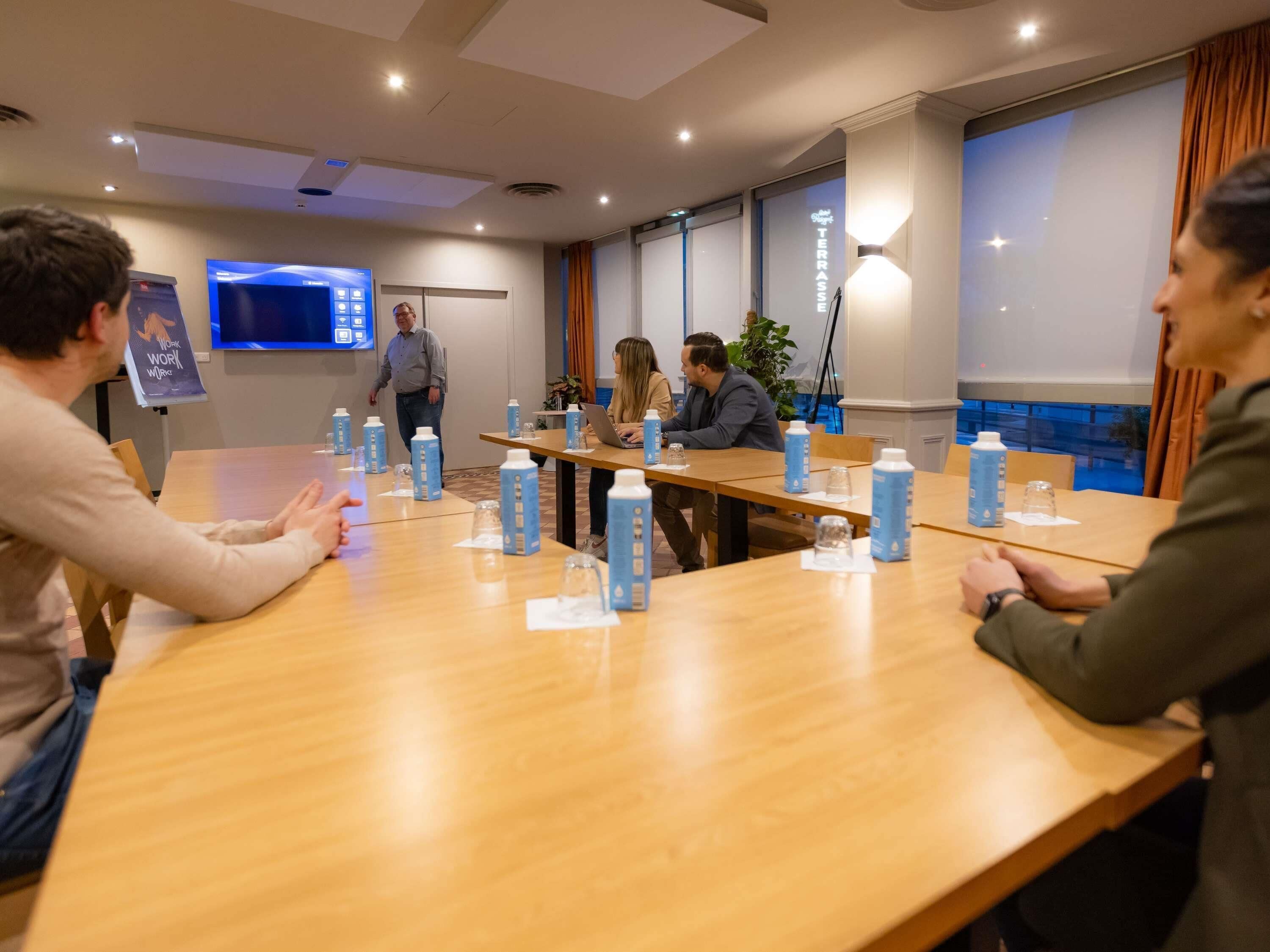A conference room with a long table, chairs, a TV screen, and a window with curtains.