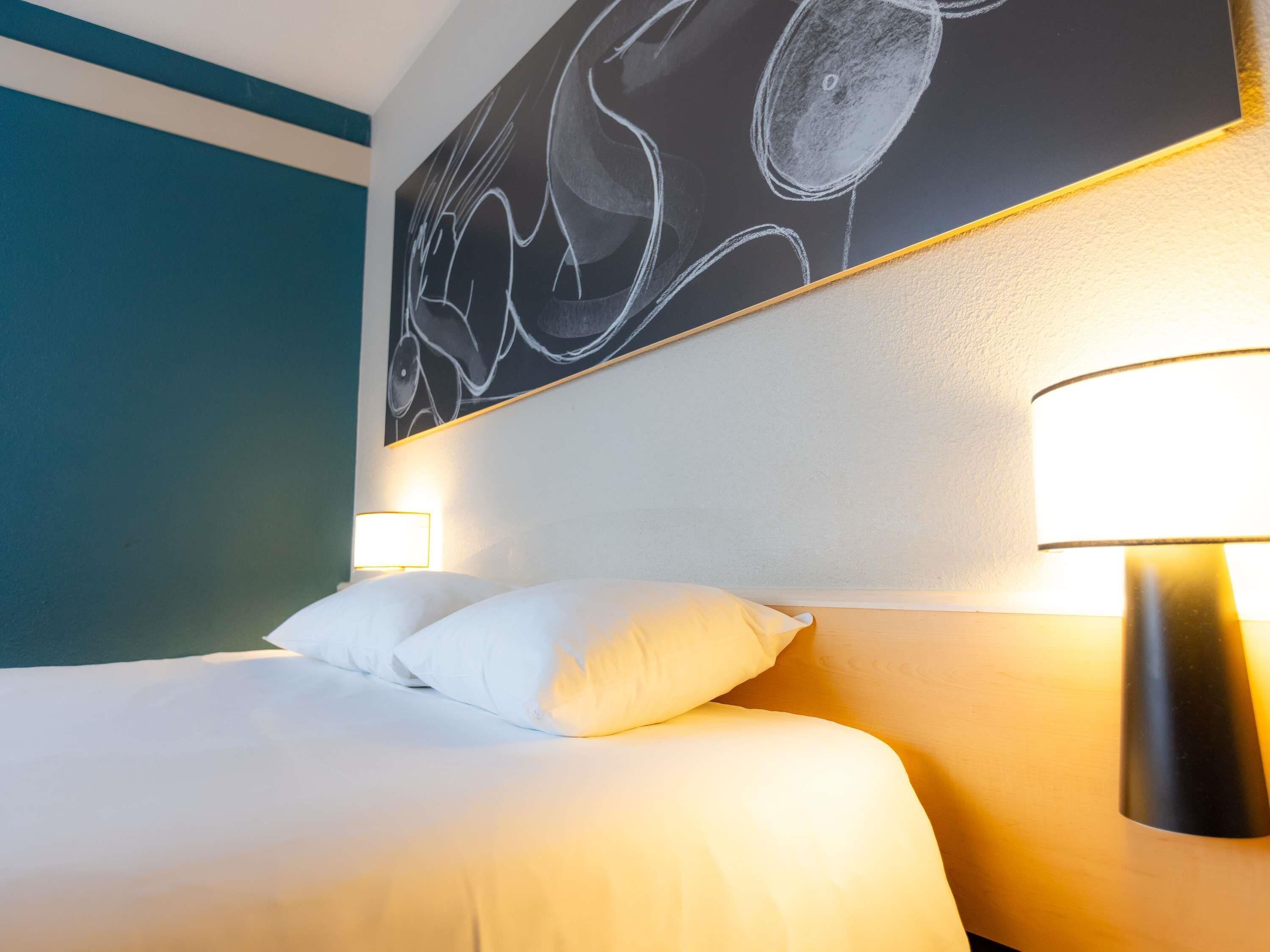 A hotel room with a bed, white pillows, a wall-mounted artwork, and a bedside lamp.