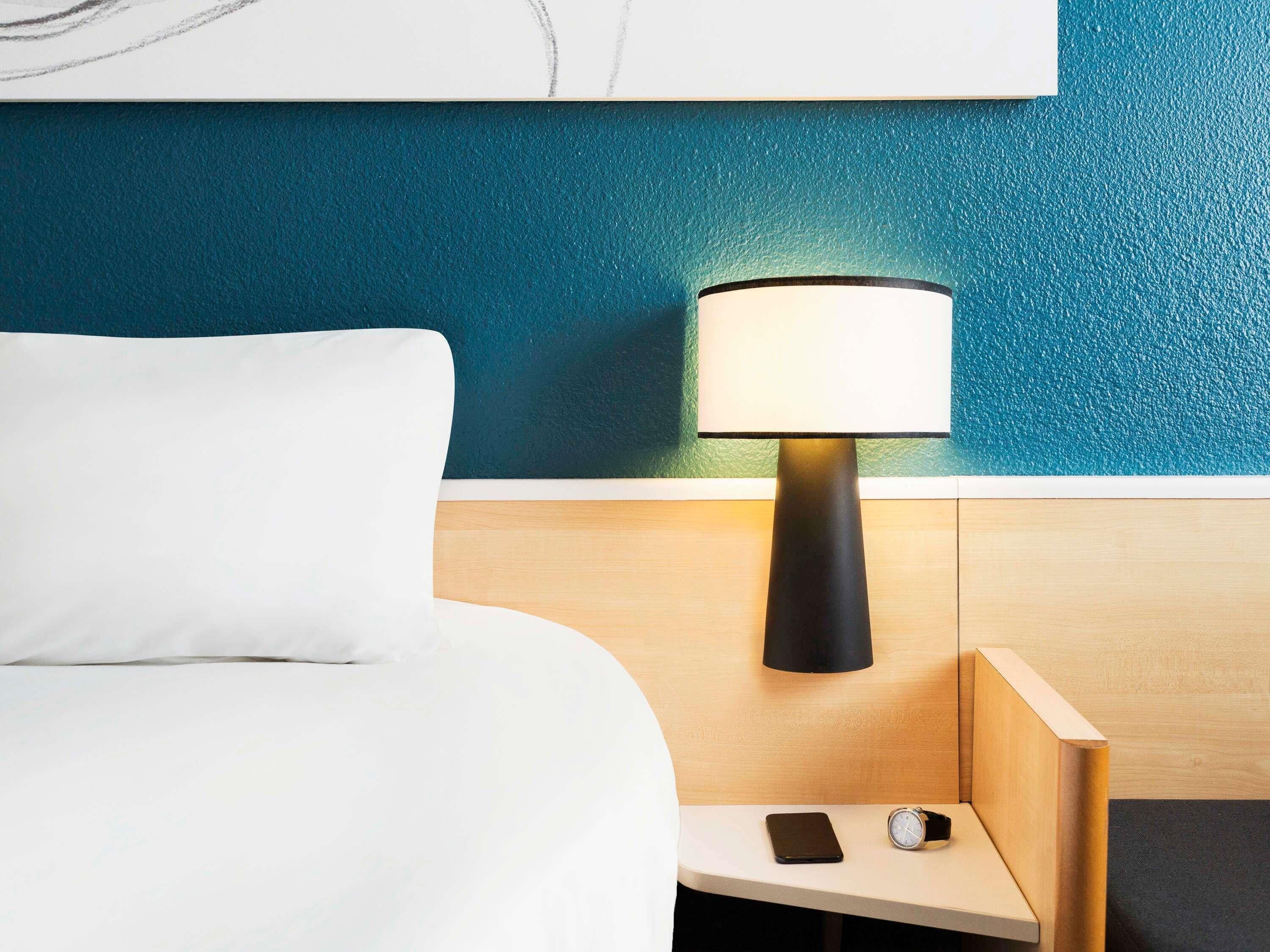 A hotel room with a bed, a nightstand, a lamp, a phone, and a watch.