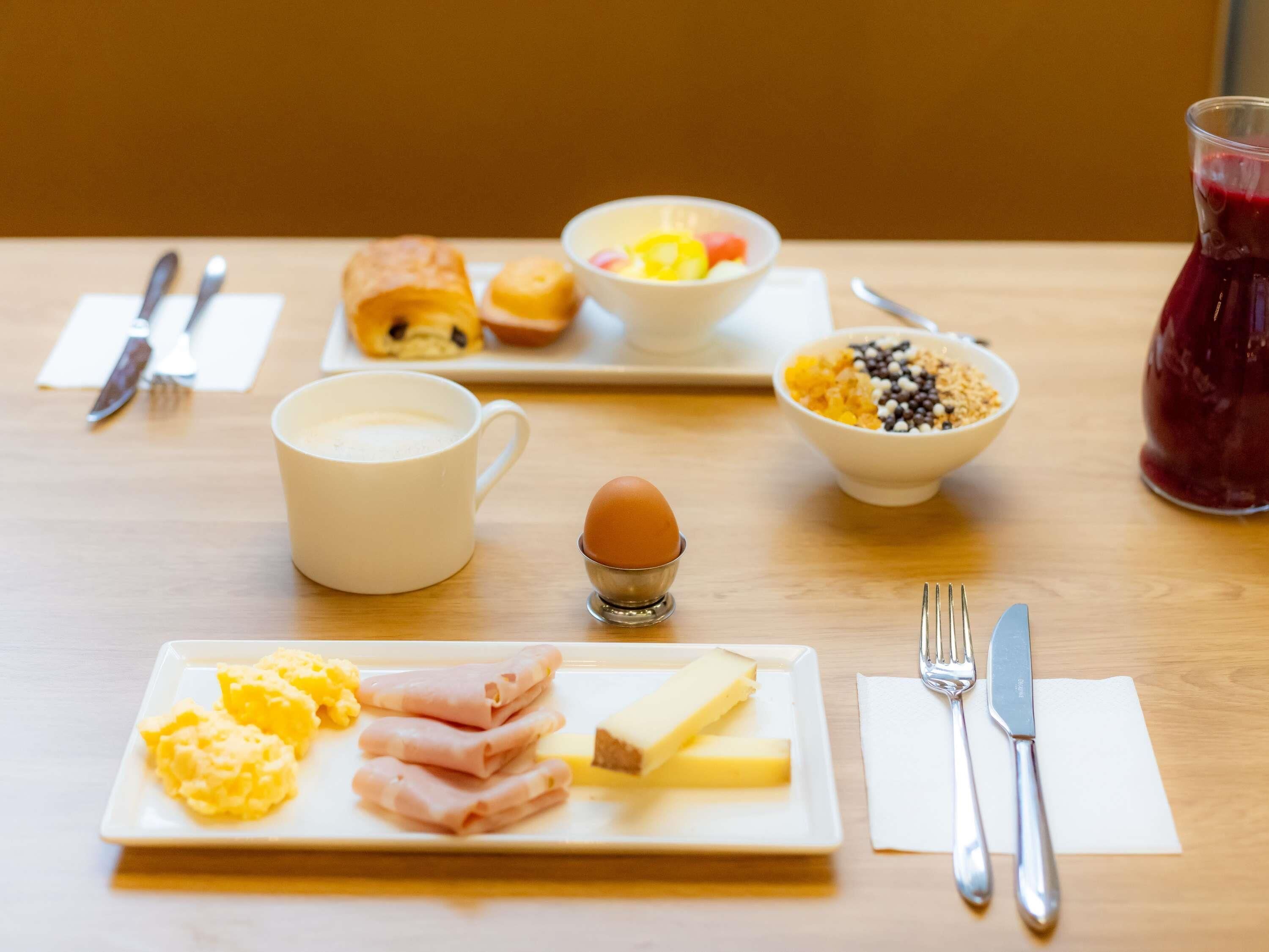 Une table de petit-déjeuner avec une assiette d’œufs, de jambon et de fromage, un bol de céréales, une tasse de café et un verre de jus de fruits.