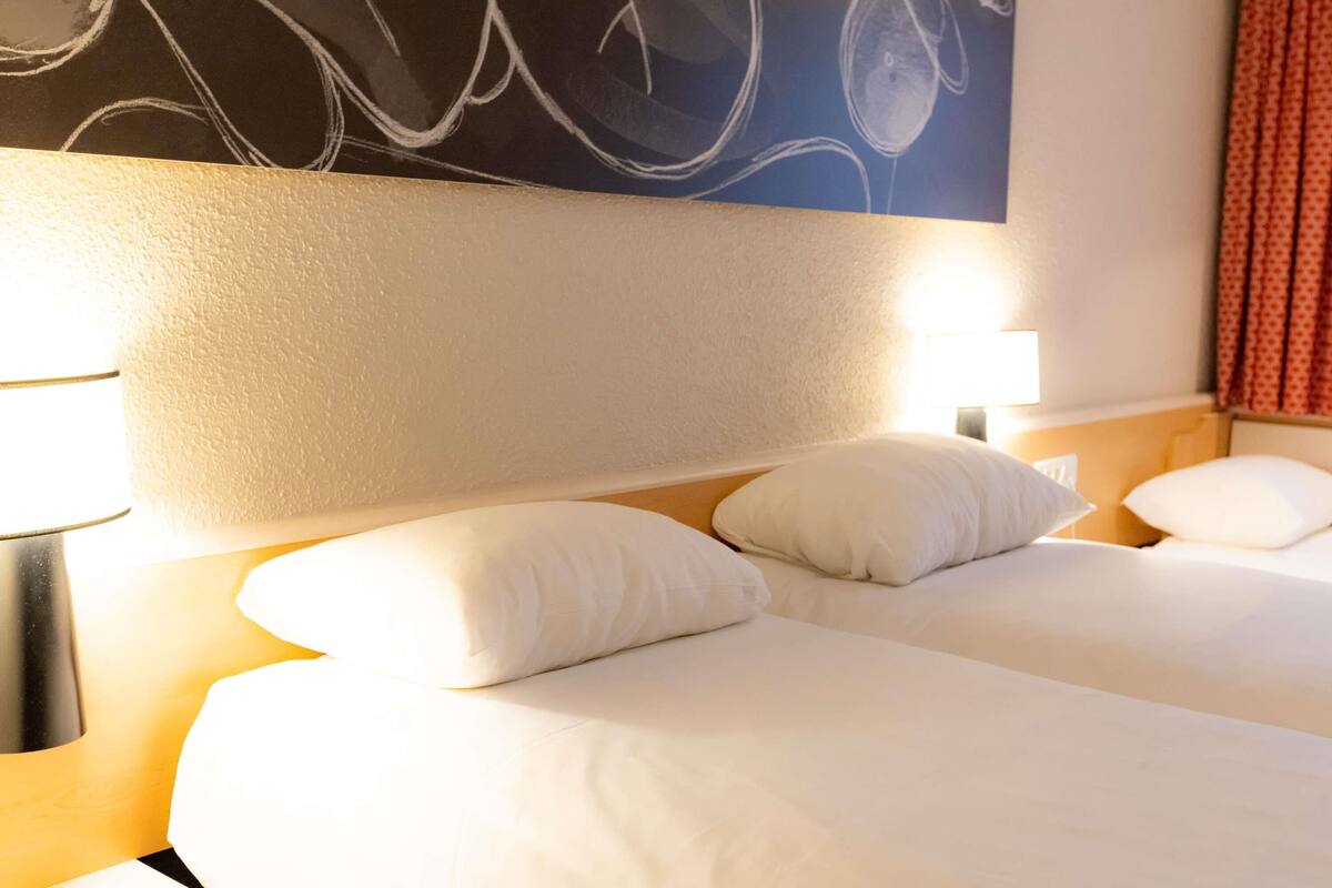 Ein Hotelzimmer mit zwei Betten, weißer Bettwäsche, einem großen abstrakten Gemälde an der Wand und einem roten Vorhang.