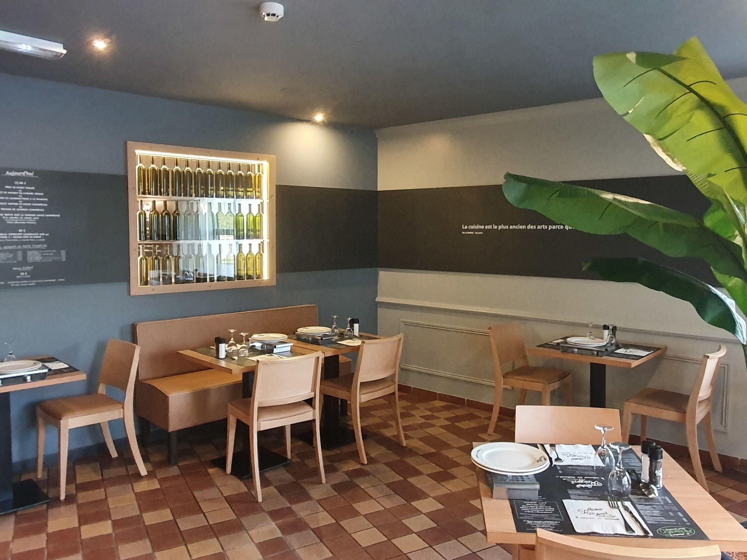 Ein Restaurantinneres mit Schachbrettboden, Holzmöbeln, einer Wandvitrine für Wein und einer großen Pflanze.