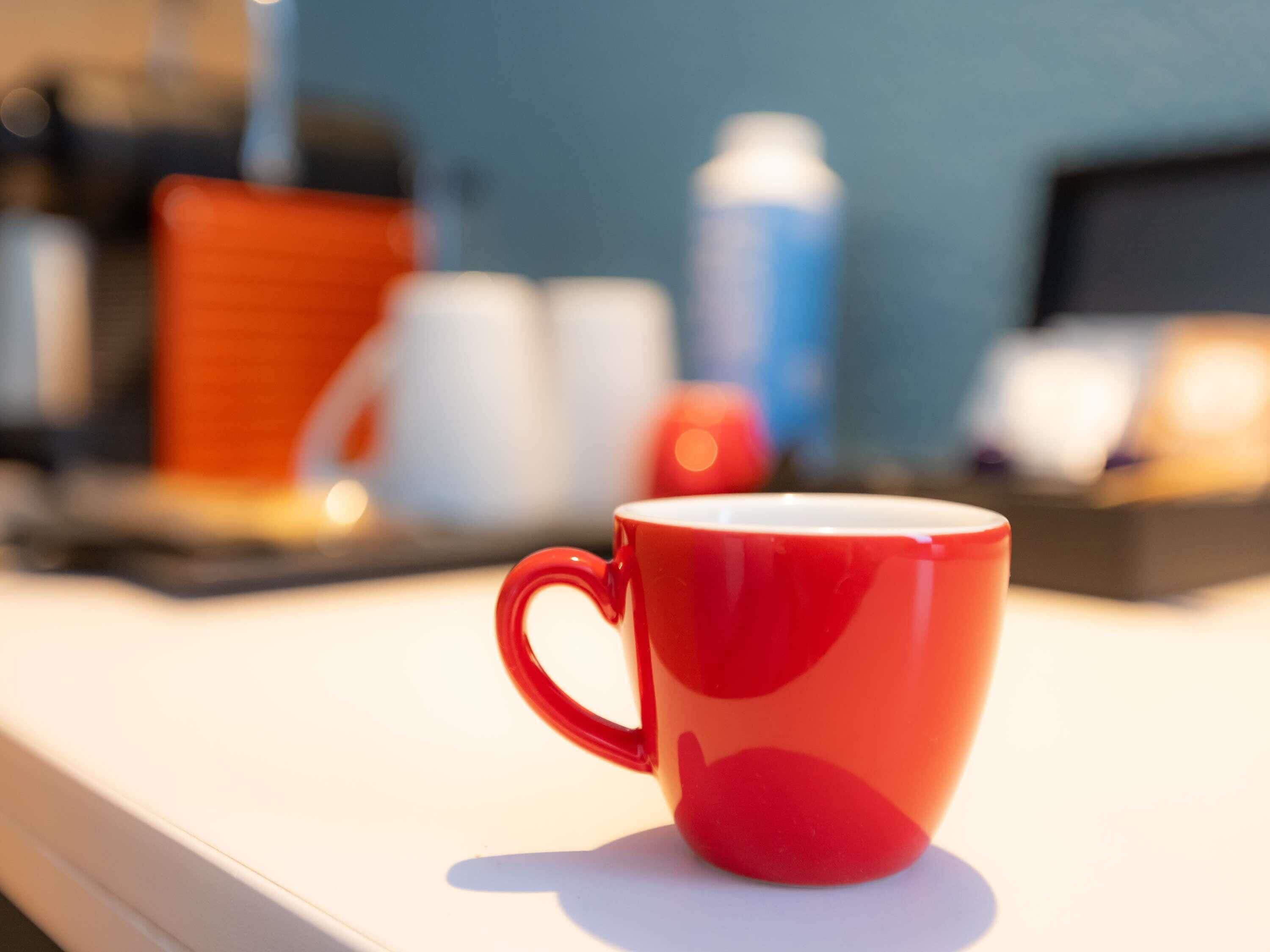 A red mug on a white surface with a blurred background of various objects.