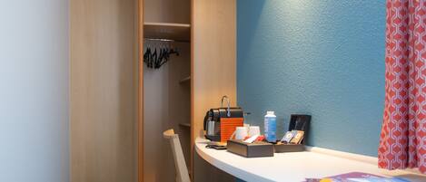 A compact room with a wooden wardrobe, a desk with a coffee maker, and a chair.