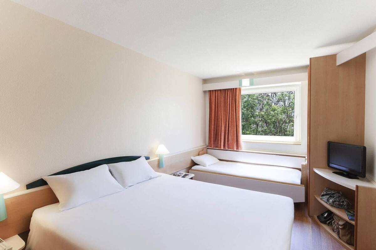 A hotel room with a large bed, a TV, a window with curtains, and a bedside table with a lamp.