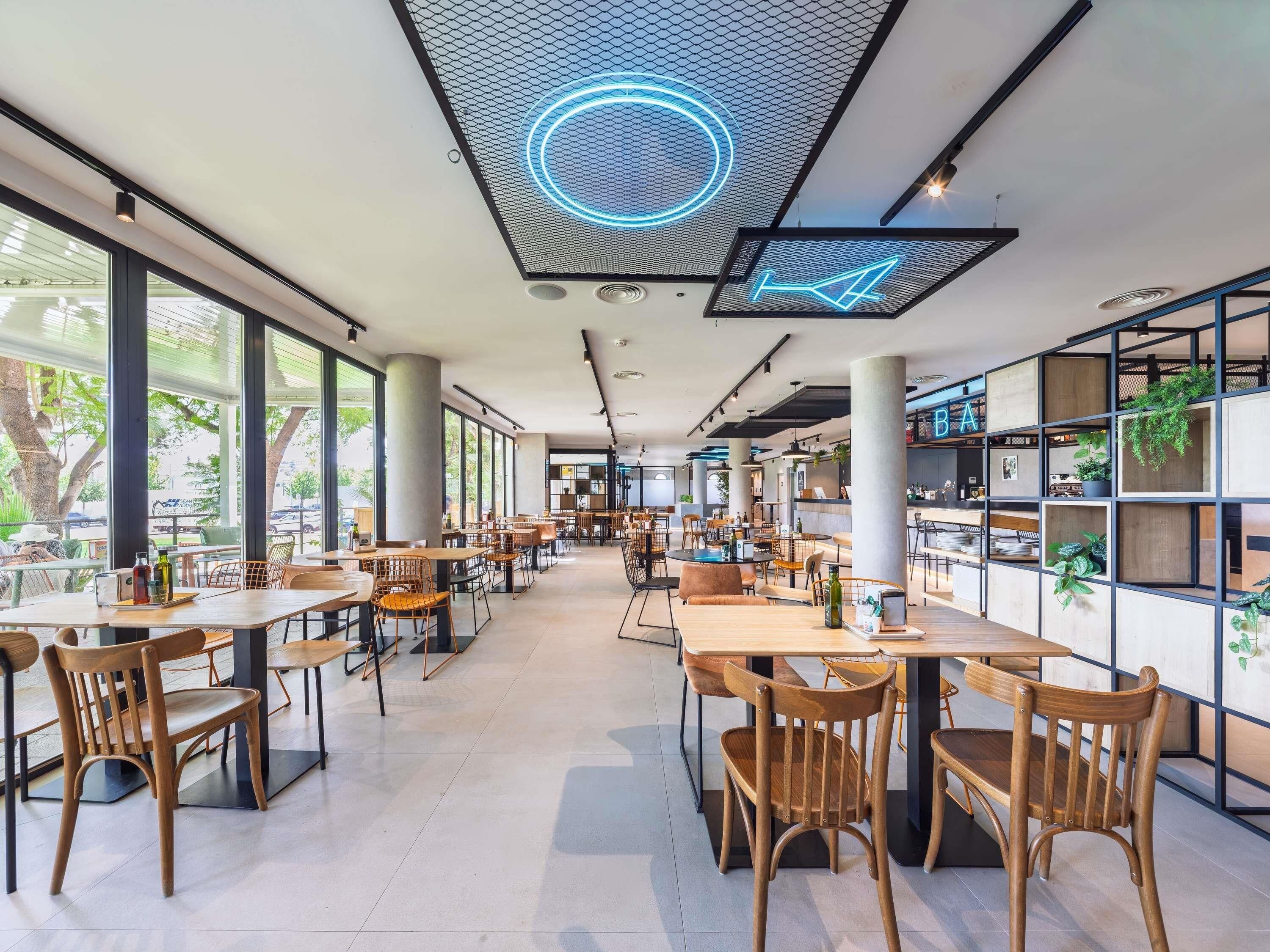 Interior moderno de una cafetería con techos altos, amplias ventanas y diversas opciones de asientos, incluyendo mesas y sillas.