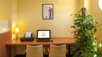 A hotel room with a desk, two chairs, a laptop, a lamp, a framed poster, and a potted plant.