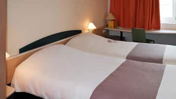A hotel room with two beds, a desk with a lamp, a chair, and a window with curtains.