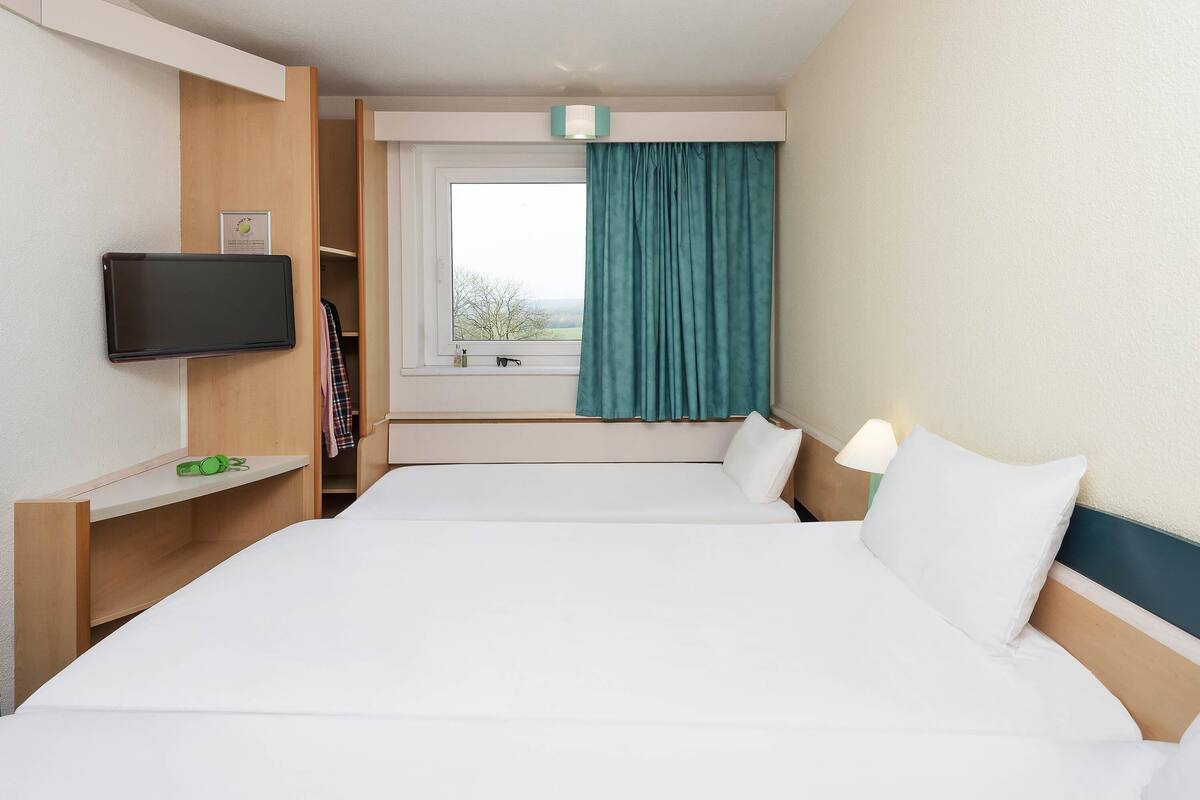Une chambre d’hôtel avec un lit, un téléviseur fixé au mur, une fenêtre avec des rideaux et une table de chevet avec une lampe.