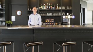 Una zona de bar con un cantinero, estantes llenos de botellas, vasos y una caja registradora.