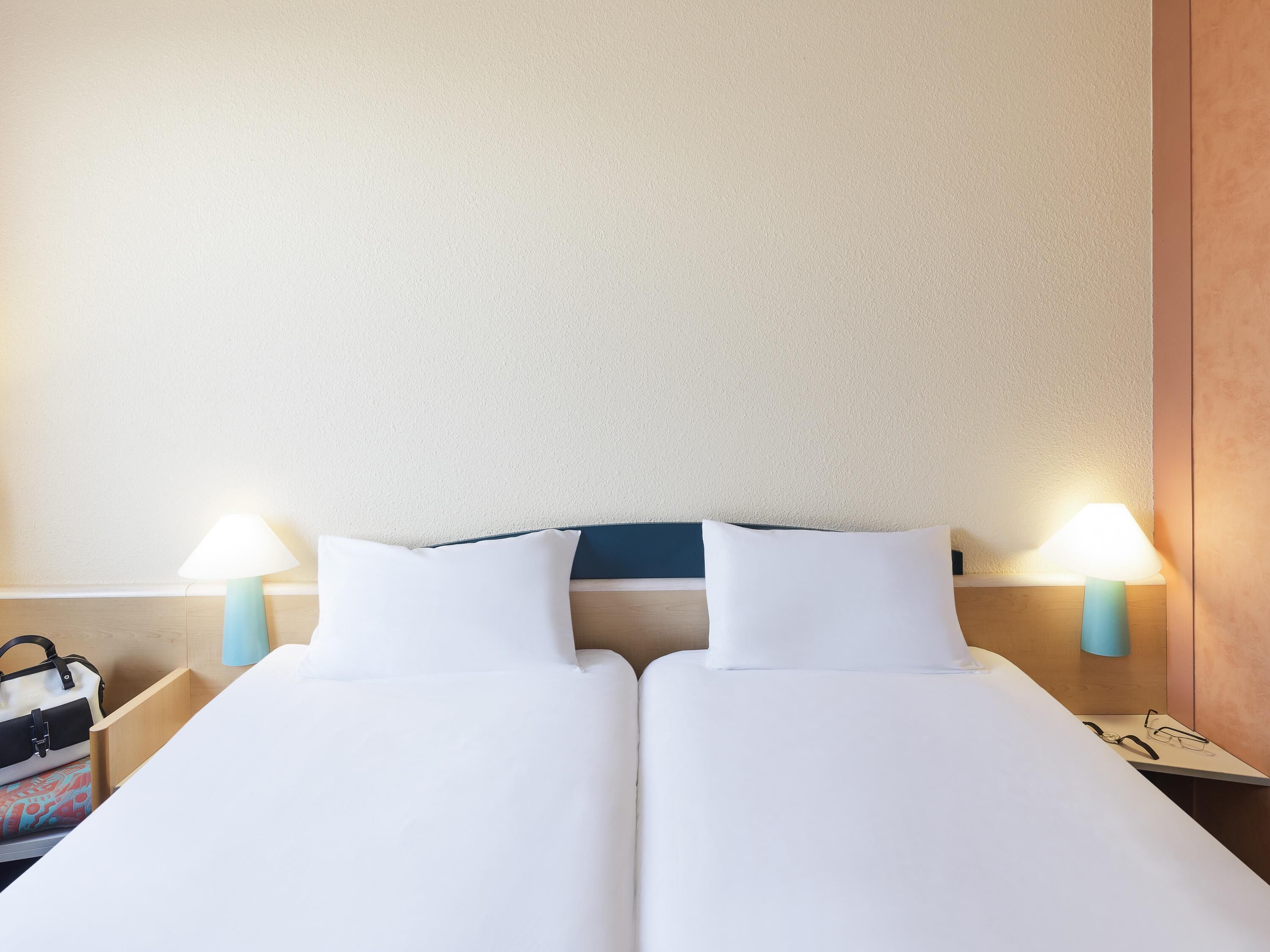 Een tweepersoonsbed met witte beddengoed, twee bedlampjes, een houten nachtkastje en een handtas aan de linkerkant.