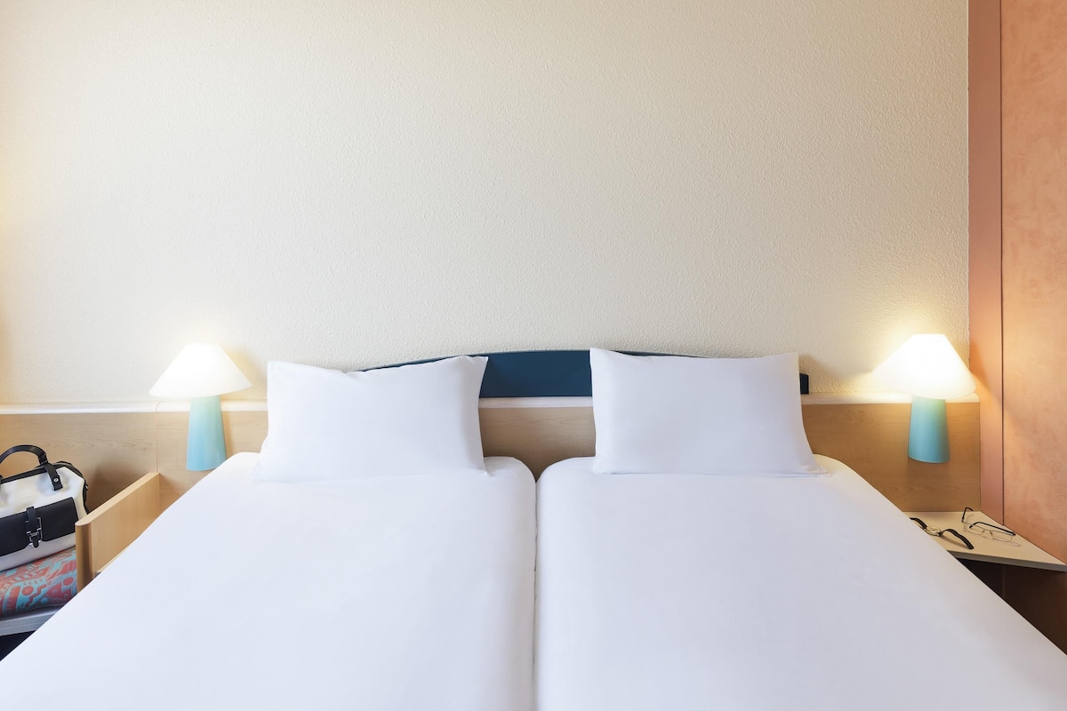 Una cama doble con ropa de cama blanca, dos lámparas de noche, un mesita de noche de madera y un bolso en el lado izquierdo.