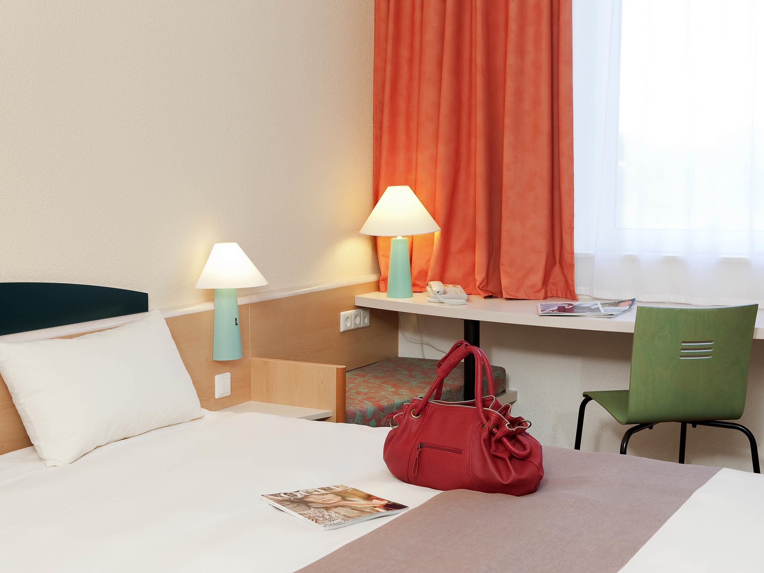 A hotel room with a bed, two bedside lamps, a desk with a chair, a red handbag, and a window with orange curtains.