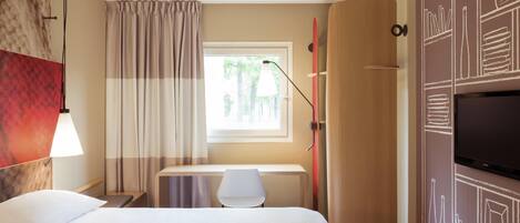 Habitación de hotel con cama, escritorio, silla, televisor y ventana con cortinas.