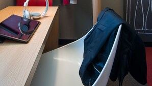 A hotel room with a wooden desk, a red wall, a white chair, a coat rack with two jackets, and a wall art piece. - ibis Manosque Cadarache (Manosque)