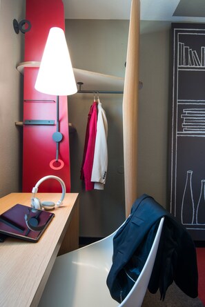 A hotel room with a wooden desk, a red wall, a white chair, a coat rack with two jackets, and a wall art piece. - ibis Manosque Cadarache (Manosque)