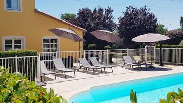 A pool area with lounge chairs and umbrellas, surrounded by a white fence and greenery.