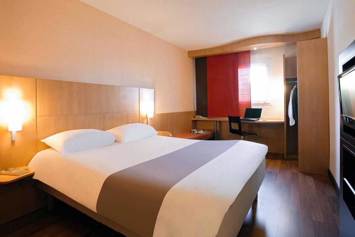 A hotel room with a bed, bedside lamps, a desk with a laptop, and a TV.
