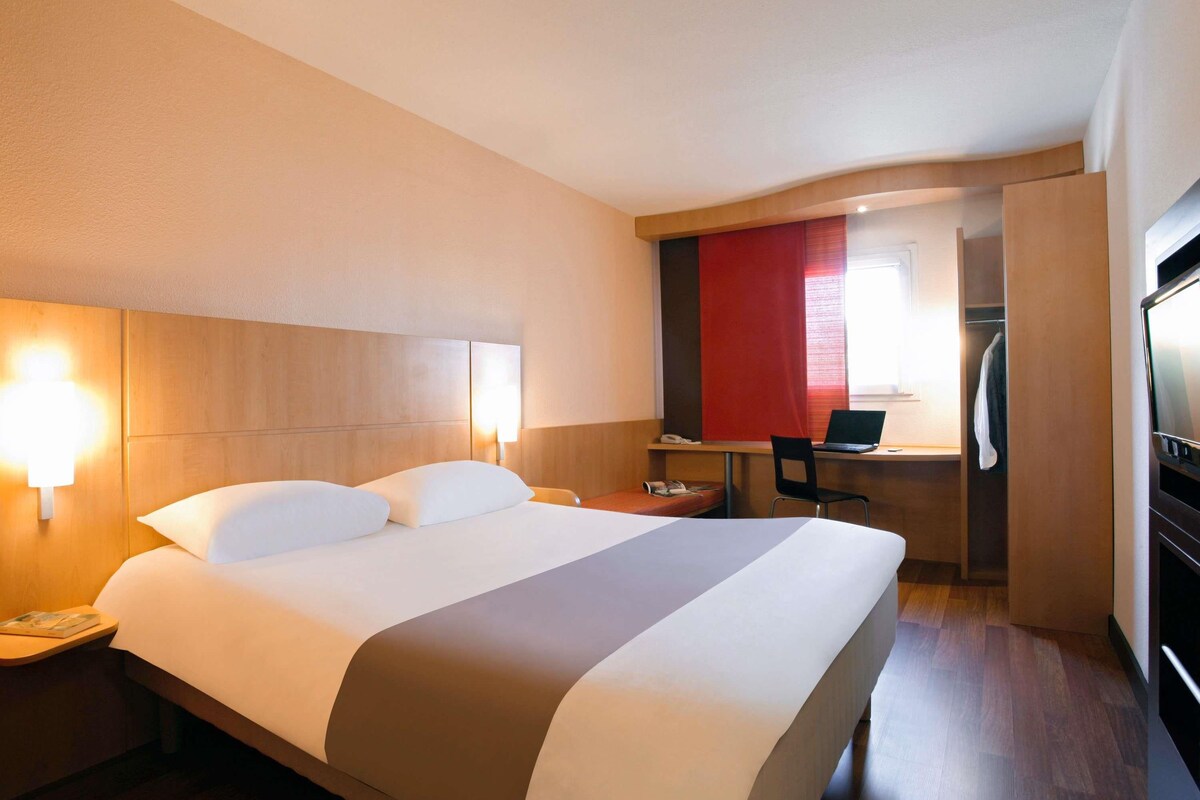 A hotel room with a bed, bedside lamps, a desk with a laptop, and a TV.