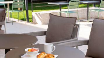 Un espace extérieur aménagé avec des tables et des chaises rondes, un saladier rempli de fruits, un croissant et une tasse blanche.
