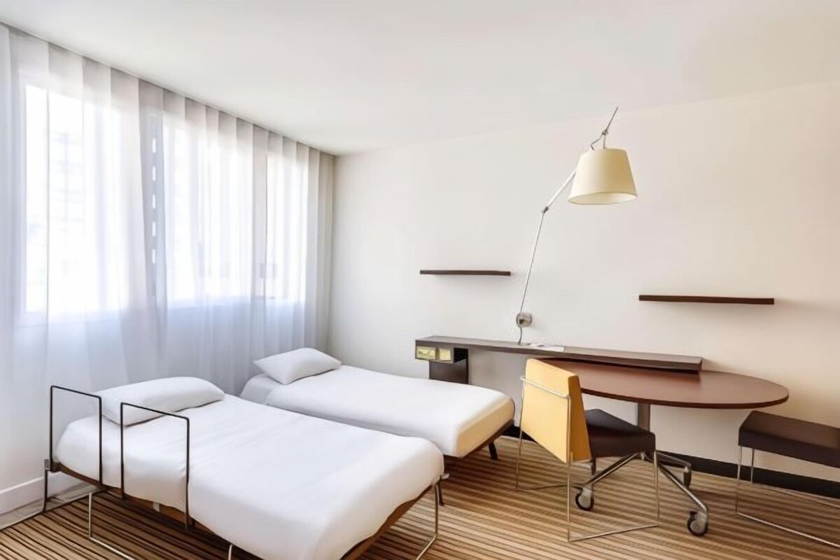 Ett hotellrum med två sängar, ett skrivbord, en stol, en lampa och ett fönster med gardiner.