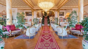Un salón amplio con columnas ornamentales, un candelabro y una alfombra roja que conduce a una escalera.