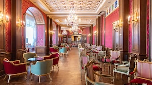 Un amplio vestíbulo de hotel con candelabros ornamentados, mobiliario rojo y dorado, y un largo pasillo que conduce a una zona de bar.