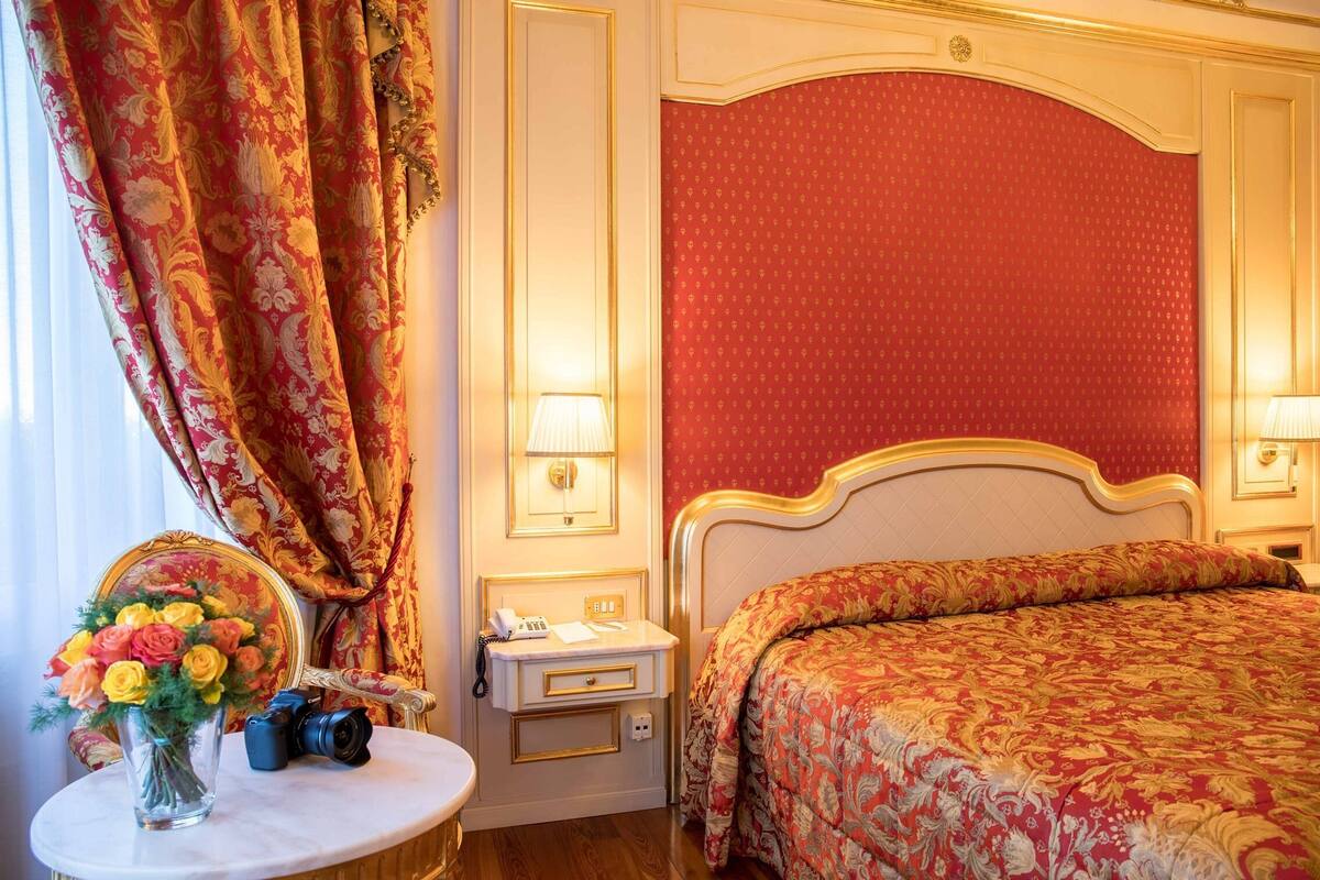 Une chambre d’hôtel avec un lit, une table de chevet avec une lampe, un miroir et un vase avec des fleurs.