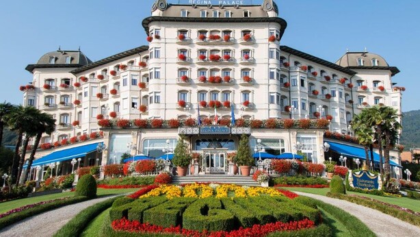 Un edificio grande de varios pisos con un estilo arquitectónico europeo clásico, con balcones adornados con macetas rojas de flores, una entrada central con un letrero y un jardín bien cuidado con diversas plantas y flores.