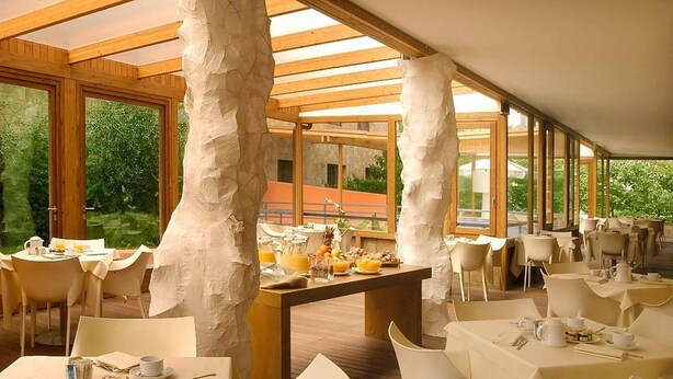 Een restaurant met een modern design, grote ramen en een houten plafond. Tafels zijn gedekt met witte tafellinnen en stoelen, en er is een buffet met eten en drinken.