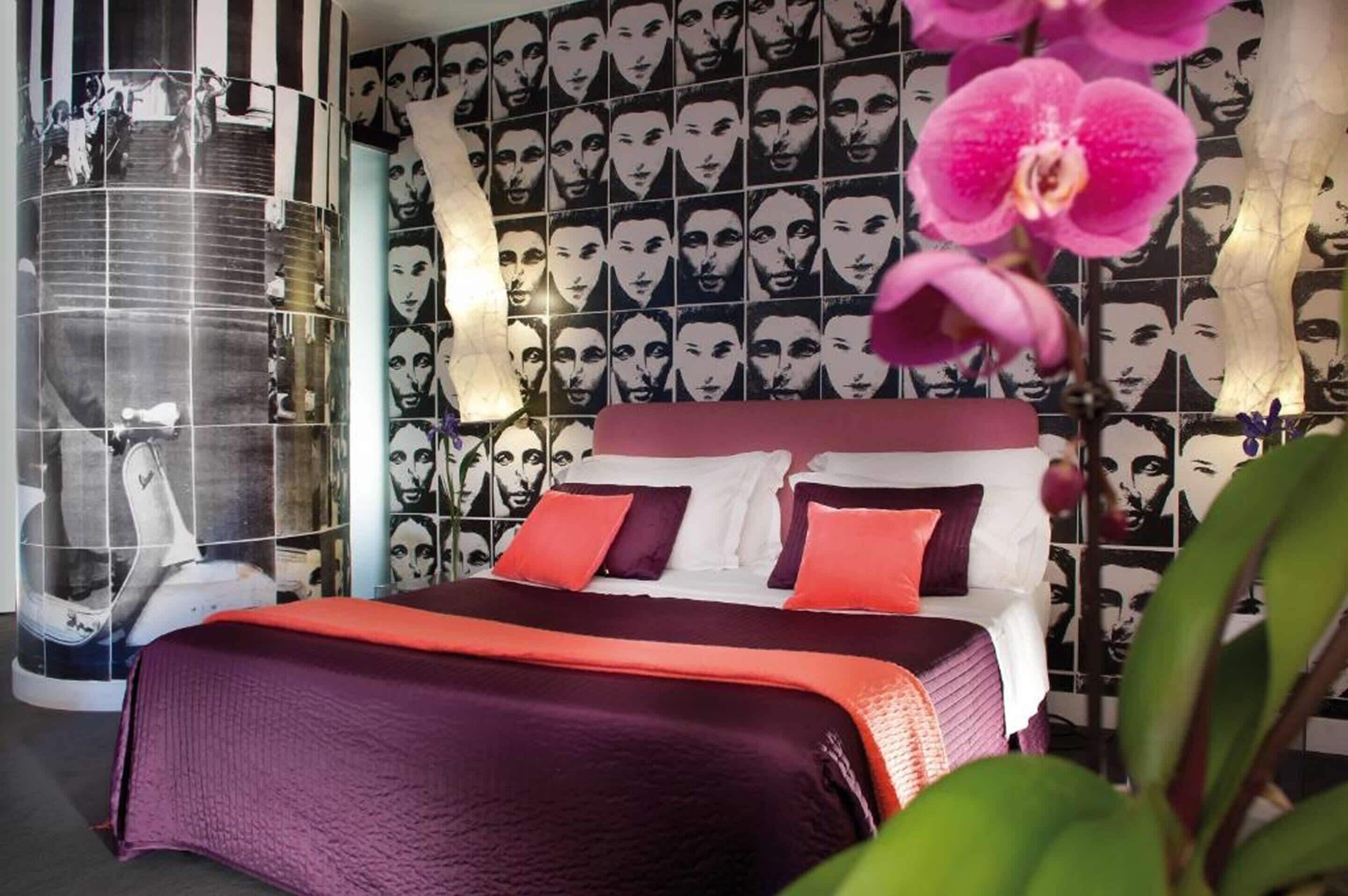 Quarto com cama, parede com imagens repetidas e uma estrutura cilíndrica grande.
