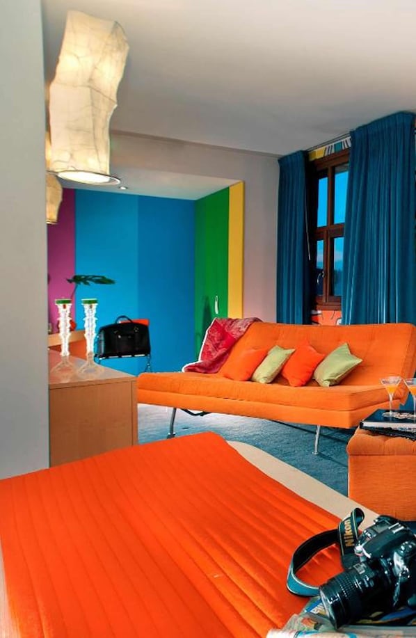 Un salon aux couleurs vives, avec un canapé orange, une fenêtre bleue et un sac noir posé sur une table d’appoint.