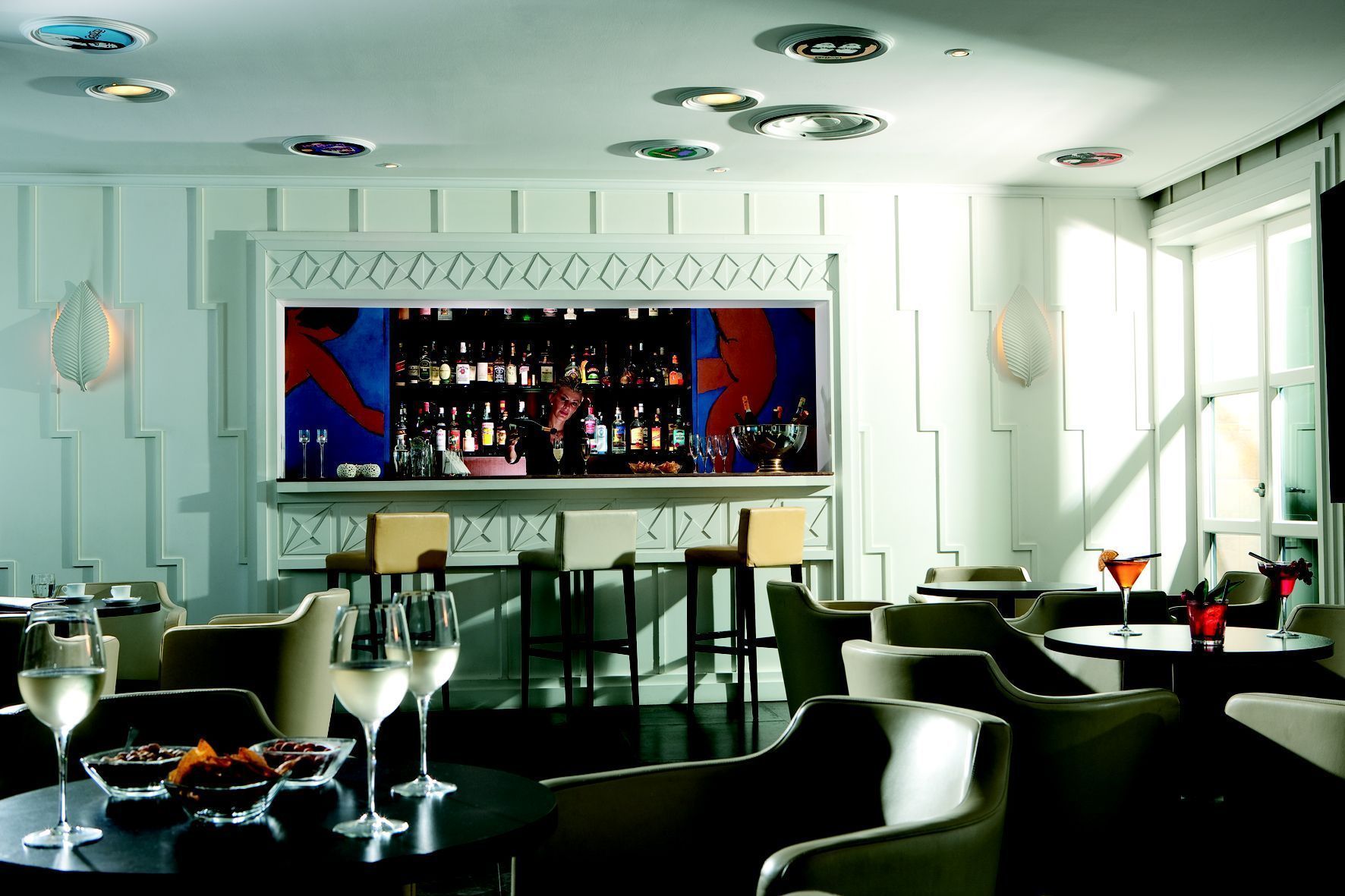 Zona de bar con un barman, una estantería repleta de licores y asientos para los clientes.
